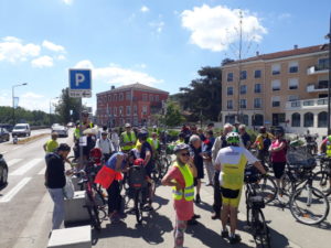 Un groupe d'une centaine de cyclistes patiente sur les abords d'une avenue. Le ciel est radieux et les contrastes dû au soleil sont prononcés.