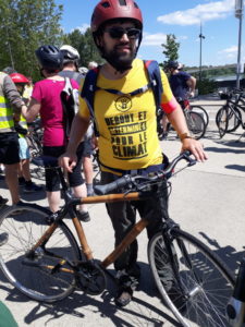 Un cycliste debout de plein pied, vélo et casque marron, montre son maillot jaune avec l'inscription noir en toutes capitales : « Debout et déterminé pour le Climat ». En arrière plan un groupe de cyclistes discute.