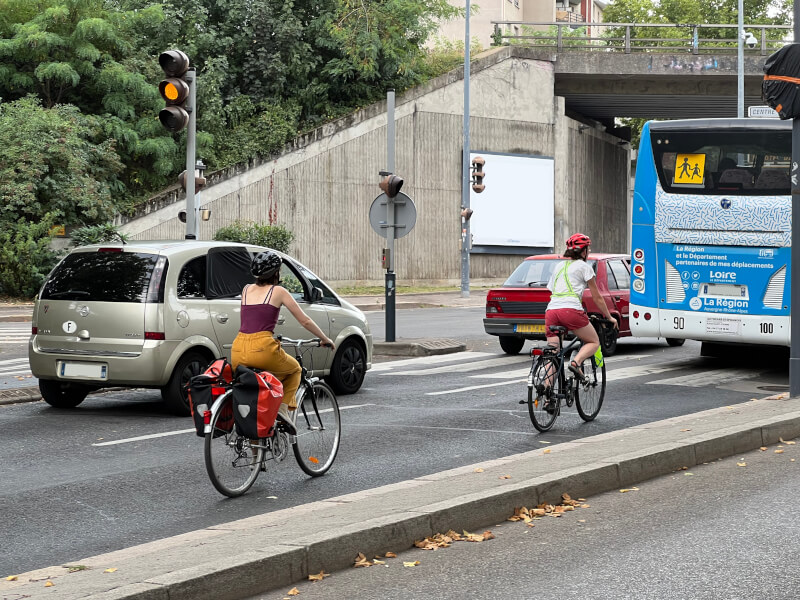 Dans la circulation auto-mobile à Saint-Étienne, deux femmes à vélo dont une avec deux sacoches arrières.