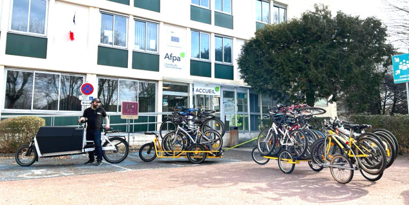 Photo devant le bâtiment de l'Afpa de Saint-Étienne. Grâce à un vélo cargo électrique, un salarié d'Ocivélo tracte trois remorques remplies de vélo : soit une vingtaine de vélos.